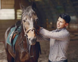 Jovem rapaz sorridente em pé ao lado do cavalo em espaço de treino com as mãos no pescoço do animal.