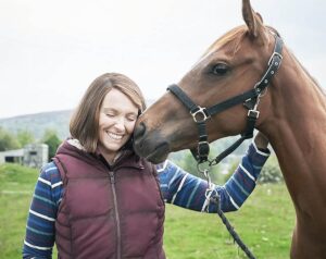 Cavalo encosta cabeça carinhosamente no rosto de uma sorridente Toni Collette de olhos fechados em um campo verde