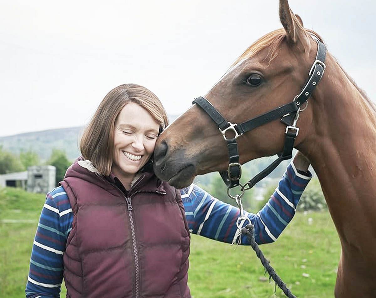 Cavalo encosta cabeça carinhosamente no rosto de uma sorridente Toni Collette de olhos fechados em um campo verde